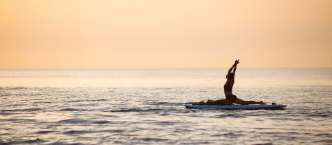 Eine Person praktiziert Yoga auf einem Paddleboard im ruhigen Ozeanwasser während eines heiteren Sonnenuntergangs, wobei der Himmel warme Orange- und Gelbtöne wirft.