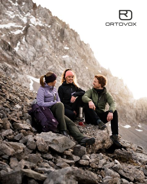 Drei Personen in Outdoor-Ausrüstung sitzen lächelnd und unterhalten sich auf felsigem Gelände in einer Bergregion. Die Sonne versinkt hinter den Gipfeln. Ein Rucksack und eine Thermoskanne stehen in der Nähe. Das Logo von Ortovox befindet sich oben rechts.