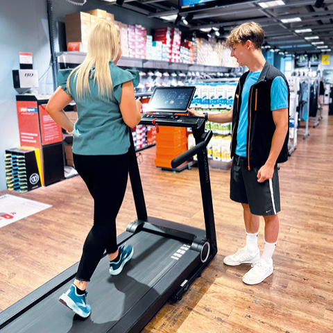Eine blonde Person läuft auf einem Laufband in einem Sportgeschäft. Ein Mitarbeiter in blauem Hemd und schwarzer Weste bedient die Konsole des Laufbands. Im Hintergrund sind Regale mit Schuhen und Sportflaschen zu sehen.