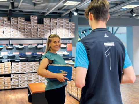 Eine Frau mit einem Schuhkarton lächelt einen Mitarbeiter in einem Sportbekleidungsgeschäft an. Im Hintergrund sind Regale voller Schuhkartons. Die Szene ist gut beleuchtet und schafft eine entspannte Einkaufsatmosphäre.