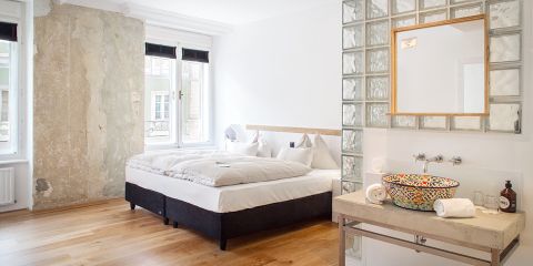 Ein minimalistisches Schlafzimmer mit Doppelbett, weißer Bettwäsche und zwei Fenstern. Das Zimmer besticht durch eine rustikale, teilweise verputzte Wand und einen Holzboden. Eine Glasbaustein-Trennwand gibt den Blick auf ein farbenfrohes Keramikwaschbecken mit einem Spiegel darüber frei.