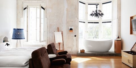 Ein helles Zimmer mit rustikalen Wänden bietet ein gemütliches Bett, zwei braune Stühle und eine freistehende Badewanne neben großen Erkerfenstern. Eine dekorative Lampe, eine Staffelei und ein Flachbildfernseher runden den Raum ab und lassen viel natürliches Licht herein.