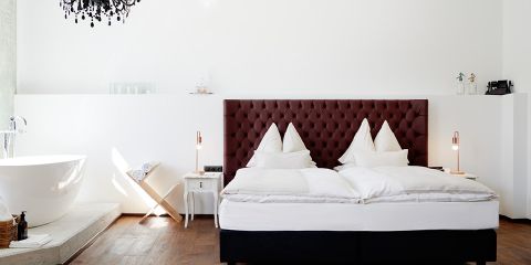 Modernes Schlafzimmer mit dunkel gepolstertem Kopfteil, weißer Bettwäsche und freistehender Badewanne. Ein schwarzer Kronleuchter hängt von der Decke. Ein kleiner Beistelltisch mit Lampen und ein Holzboden verleihen dem minimalistischen Design Wärme.