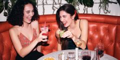 Zwei Frauen sitzen lächelnd auf einem roten Sofa in einer Bar und stoßen mit Cocktailgläsern an. Auf dem Tisch vor ihnen stehen Getränke, Snacks und eine Kerze. Hängepflanzen schmücken den Hintergrund.