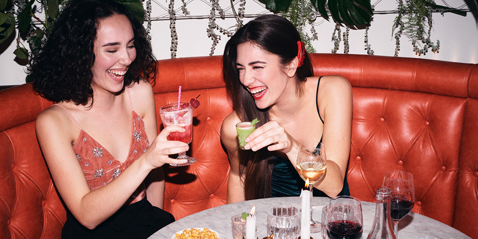 Zwei Frauen sitzen lächelnd auf einem roten Sofa in einer Bar und stoßen mit Cocktailgläsern an. Auf dem Tisch vor ihnen stehen Getränke, Snacks und eine Kerze. Hängepflanzen schmücken den Hintergrund.