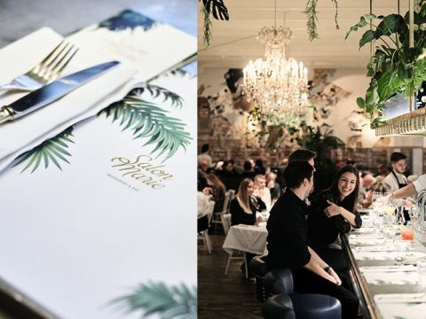 Nahaufnahme einer Speisekarte mit Palmblatt-Design auf der linken Seite. Auf der rechten Seite sitzen Menschen an einer Bar in einem eleganten Restaurant mit Kronleuchtern und Grünpflanzen, unterhalten sich und genießen Getränke.