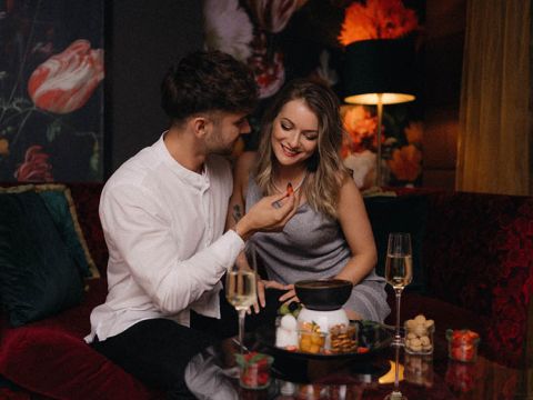Ein Paar sitzt eng aneinander gekuschelt auf einem gemütlichen, blumengeschmückten Sofa. Der Mann bietet der Frau mit einem liebevollen Lächeln Schokolade an. Zwei Gläser Champagner und ein Fondue-Set mit Snacks stehen auf dem Tisch. Gedämpftes Licht schafft eine intime Atmosphäre.