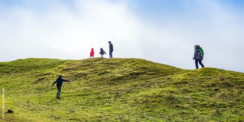 Eine Gruppe von Menschen, darunter ein Kind in einer roten Jacke, geht unter strahlend blauem Himmel auf einem grasbewachsenen Hügel spazieren. Sie sind entlang des Hangs verstreut, einige klettern hinauf, andere gehen oben entlang. Die Landschaft ist lebendig und offen.