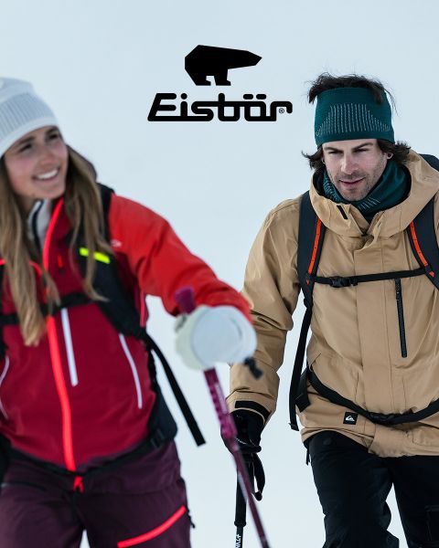 Eine Frau und ein Mann lächeln beim Skifahren im verschneiten Gelände. Sie trägt eine rote Jacke und eine weiße Mütze und hält Skistöcke in der Hand, er eine beige Jacke und eine grüne Mütze. Über ihnen prangt das „Eisbär“-Logo.