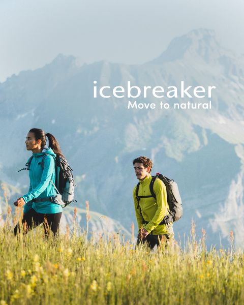 Zwei Personen wandern in einer Berglandschaft. Sie tragen Rucksäcke und Outdoor-Kleidung. Auf dem Bild ist der Schriftzug „Icebreaker Move to Natural“ zu sehen. Gelbe Wildblumen sind im Vordergrund zu sehen, im Hintergrund neblige Berge.