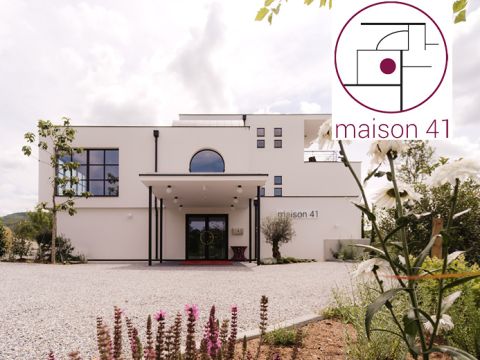 Hinter einer Kiesauffahrt steht ein modernes, weißes zweistöckiges Gebäude mit großen Fenstern und der Aufschrift „Maison 41“. Im Vordergrund blühen Pflanzen. Das Logo im Hintergrund zeigt abstrakte geometrische Formen und den Text „Maison 41“.