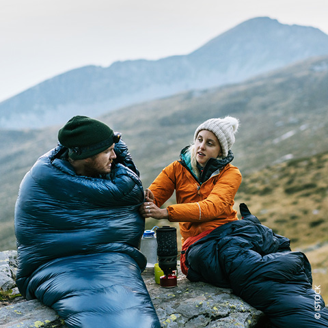 Zwei Personen sitzen im Freien auf einem felsigen Untergrund, eingehüllt in Schlafsäcke. Sie tragen warme Kleidung, darunter Mützen und Jacken. Ein Rucksack und ein Campingkocher stehen in der Nähe. Im Hintergrund sind Berge und ein bewölkter Himmel zu sehen.