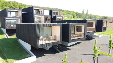 Moderne schwarze, würfelförmige Häuser mit großen Fenstern und Terrassen liegen an einem grünen Hang. Jede Wohneinheit ist erhöht und verfügt über eine Treppe zum Eingang. Im Vordergrund sind Bäume gepflanzt, im Hintergrund ist ein Waldgebiet zu sehen.