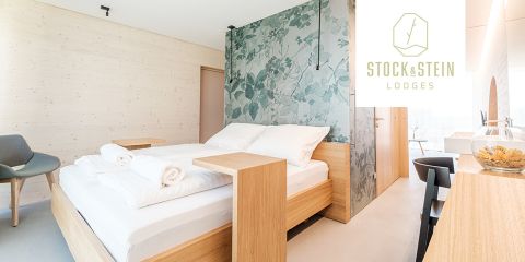 Modernes Hotelzimmer mit Holzbett, weißer Bettwäsche und gerollten Handtüchern. Mit floraler Akzentwand und hellen Holzmöbeln. Ein grauer Stuhl und eine Glasschiebetür sind sichtbar. Das Logo der Stock & Stein Lodges befindet sich in der oberen rechten Ecke.