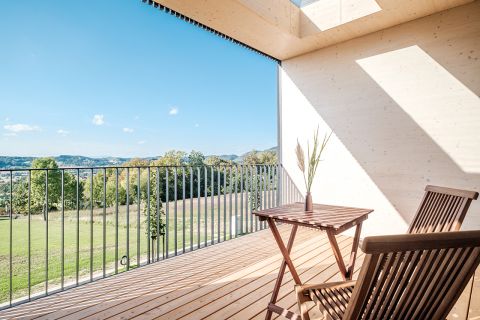 Ein Holzbalkon mit Tisch und Stühlen und Geländer bietet Aussicht auf eine üppige grüne Wiese und ferne Hügel unter einem klaren blauen Himmel. Ein Oberlicht wirft Sonnenlicht auf den Holzboden.