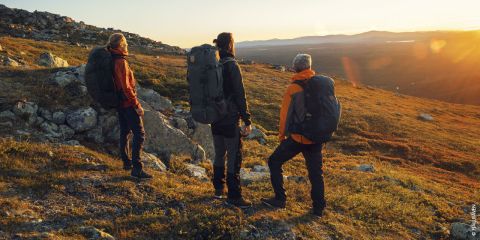 Drei Wanderer mit Rucksäcken stehen auf einem felsigen Hügel und genießen den malerischen Sonnenuntergang über fernen Bergen und einem See. Sie tragen Outdoor-Ausrüstung und sind von spärlicher Vegetation umgeben, während die untergehende Sonne warm und golden leuchtet.