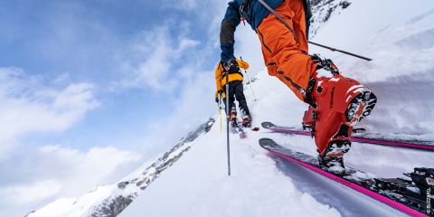 Zwei Skifahrer in leuchtender Ausrüstung fahren mit Stöcken einen verschneiten Hang hinauf. Einer trägt orangefarbene Hosen und rote Stiefel, der andere ist etwas weiter vorne in gelben Schuhen. Im Hintergrund sind schneebedeckte Berge und ein blauer Himmel zu sehen.