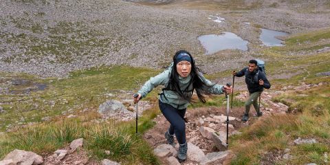 Zwei Personen wandern einen felsigen Pfad in einer Bergregion hinauf. Die Frau vorne trägt eine hellgrüne Jacke und benutzt Wanderstöcke. Der Mann dahinter folgt mit einem Rucksack. Im Hintergrund sind ein kleiner Teich und Grasflächen zu sehen.