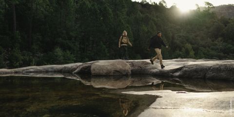 Zwei Menschen wandern auf großen Felsen neben einem spiegelnden Gewässer, umgeben von dichtem Wald. Die Sonne scheint durch die Bäume und schafft eine ruhige Naturkulisse. Sie tragen Rucksäcke, was auf eine Wanderung oder ein Abenteuer hindeutet.