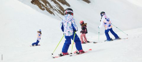 Kinder in farbenfroher Winterkleidung und mit Helmen fahren auf einem verschneiten Hang Ski. Sie stehen mit Abstand zueinander und halten jeweils einen Skistock in der Hand, während sie den Hang hinuntergleiten. Im Hintergrund ist unter klarem Himmel ein felsiger, schneebedeckter Berg zu sehen.