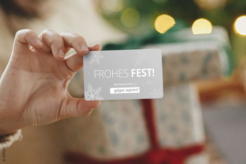 Eine Person hält eine Karte mit der Aufschrift „Frohes Fest!“. Im Hintergrund liegt ein mit einer roten Schleife verpacktes Geschenk vor einem geschmückten Weihnachtsbaum mit leuchtenden Lichtern.