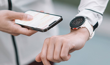Eine Person mit weißem Ärmel überprüft ihre Smartwatch, während sie ein Smartphone in der Hand hält. Die Smartwatch hat ein rundes Zifferblatt mit weißem Armband, und der Smartphone-Bildschirm ist teilweise sichtbar. Der Hintergrund ist unscharf.
