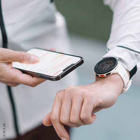 Eine Person in einem weißen Langarmshirt hält in einer Hand ein Smartphone, das Fitnessdaten anzeigt. Am anderen Handgelenk hängt eine weiße Smartwatch mit rundem Zifferblatt. Der Fokus liegt auf den Geräten vor einem neutralen Hintergrund.