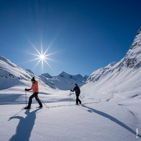 Zwei Personen wandern an einem sonnigen Tag mit Schneeschuhen durch eine verschneite Berglandschaft. Die Sonne scheint hell vom klaren blauen Himmel und wirft lange Schatten auf den Schnee. Im Hintergrund sind majestätische Gipfel zu sehen.