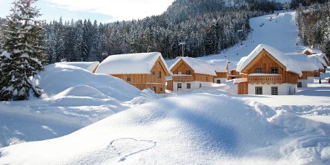 Schneebedeckte Holzhütten, umgeben von einem Wald mit schneebedeckten Bäumen. Im Hintergrund ist eine Skipiste mit Skifahrern zu sehen. Im Vordergrund ist ein Herz in den Schnee gezeichnet. Die Szene ist friedlich und winterlich.