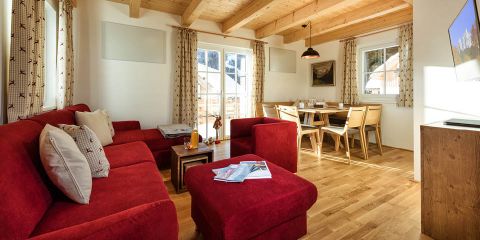 Gemütliches Wohnzimmer mit einem roten Ecksofa und passendem Ottoman auf Holzboden. Im Hintergrund steht ein Esstisch mit Stühlen neben einem Fenster mit Vorhängen. An der Wand hängt ein Flachbildfernseher, die Decke hat freiliegende Holzbalken.