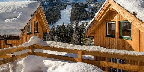 Zwei Holzhütten mit schneebedeckten Dächern liegen eingebettet in einer Winterlandschaft. Im Vordergrund ist ein Holzzaun zu sehen, im Hintergrund sind schneebedeckte Bäume und Berge unter einem klaren Himmel zu sehen.