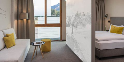 Modernes Hotelzimmer mit Bergblick. Es verfügt über ein Doppelbett mit gelben und grauen Kissen, ein Sofa, einen kleinen runden Tisch und einen gelben Ottomanen. Ein großes Fenster und Bergkunst-Dekor erhellen das grau-weiße Interieur.