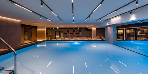 Innenpoolbereich mit elegantem, modernem Design. Der Pool ist mit sanftem Blaulicht beleuchtet und von Liegestühlen umgeben. Die Decke ist mit linearen Lichtern ausgestattet, die eine moderne Atmosphäre schaffen. Glaswände auf einer Seite führen zu einem anderen Bereich.
