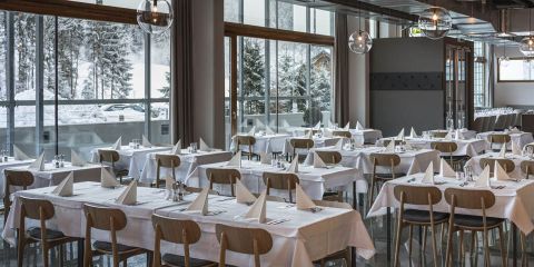 Ein geräumiges Restaurant mit großen Fenstern, die den Blick auf eine verschneite Landschaft freigeben. Die Tische sind hübsch mit weißen Tischdecken, gefalteten Servietten und Besteck gedeckt. Deckenleuchten verleihen dem modernen und eleganten Ambiente Wärme.