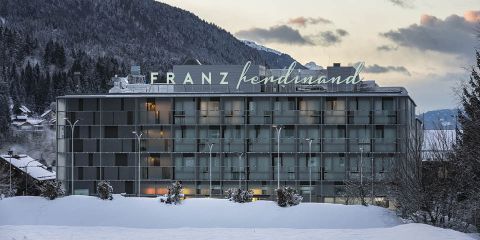 Ein modernes, mehrstöckiges Hotelgebäude mit der Aufschrift „Franz Ferdinand“ steht inmitten einer verschneiten Landschaft mit Bergen im Hintergrund. Die Glasfassade reflektiert das sanfte Licht der Dämmerung, im Vordergrund sind die Bäume leicht mit Schnee bestäubt.