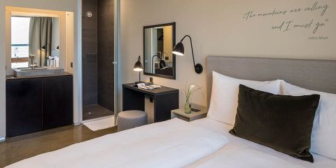 Modernes Hotelzimmer mit großem Bett, weißer Bettwäsche und dunkelgrünem Kissen. Ein kleiner Schreibtisch, eine Lampe und ein Spiegel stehen seitlich. Offenes Badezimmer mit Waschbecken und Handtüchern. An der Wand hängt ein Zitat: „Die Berge rufen, und ich muss gehen.“.