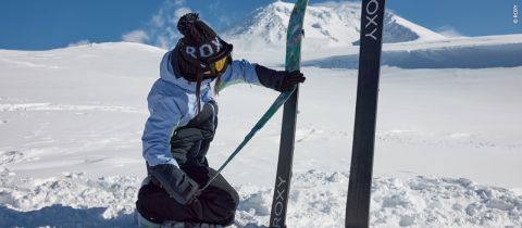 Eine Person in Winterkleidung kniet im Schnee und richtet ein Paar Skier mit dem „Roxy“-Schriftzug. Sie trägt eine schwarz-weiße Mütze, eine Skibrille und eine blaue Jacke. Im Hintergrund sind schneebedeckte Berge zu sehen.