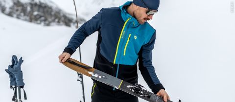 Eine Person in einer blau-schwarzen Jacke, mit Hut und Sonnenbrille richtet einen Ski auf einem verschneiten Hang aus. In der Nähe sind graue Handschuhe an Skistöcken befestigt. Im Hintergrund ist eine schneebedeckte Landschaft zu sehen.