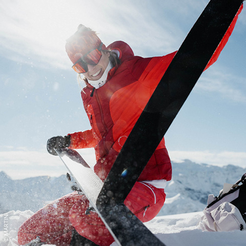 Eine Person in roter Skiausrüstung sitzt lächelnd auf einem schneebedeckten Berg und passt ihre Skiausrüstung an. Sonnenlicht fällt herab, im Hintergrund sind schneebedeckte Gipfel zu sehen.