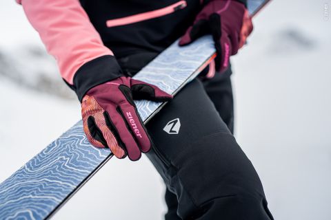 Eine Person mit rosa und burgunderfarbenen Handschuhen hält einen Ski mit blau gemusterter Oberfläche vor ihrer schwarzen Hose mit Logo. Der verschneite Hintergrund lässt an eine winterliche Umgebung denken.