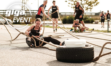 Eine Gruppe von Menschen nimmt an einem Outdoor-Fitness-Event teil. Eine lächelnde Frau sitzt auf dem Boden und zieht mit einem Seil einen Reifen. Im Hintergrund sind andere mit ähnlichen Aktivitäten beschäftigt. Das Logo trägt die Aufschrift „giga EVENT“.
