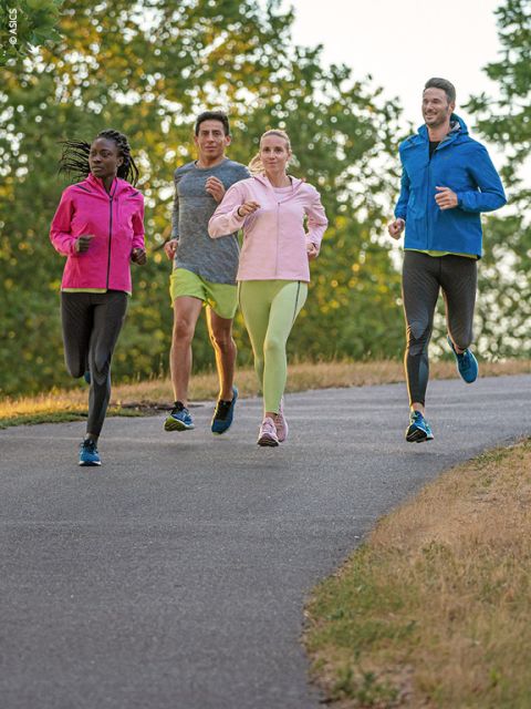 Vier Personen joggen auf einem gepflasterten Weg durch einen Park. Sie tragen Sportkleidung mit hellen Jacken und Laufschuhen. Im Hintergrund sind grüne Bäume unter einem klaren Himmel zu sehen, was einen sonnigen Tag suggeriert.