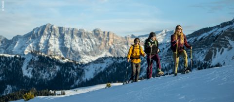 Drei Personen wandern mit Schneeschuhen einen verschneiten Hügel hinauf, im Hintergrund schneebedeckte Berge. Sie tragen Winterkleidung und Sonnenbrillen und genießen den sonnigen Tag in der malerischen Schneelandschaft.