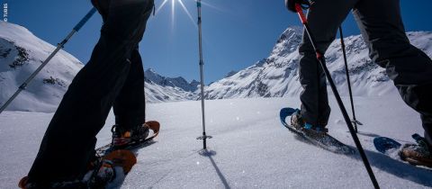 Zwei Personen wandern mit Schneeschuhen durch eine verschneite Berglandschaft unter strahlend blauem Himmel. Die Sonne scheint hell und wirft Schatten auf den unberührten Schnee. Im Hintergrund erheben sich majestätische Gipfel.