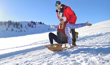 Eine Person in Winterkleidung schiebt eine andere Person auf einem Schlitten einen schneebedeckten Hang hinunter. Die Sonne scheint hell, der Himmel ist klar und blau. Beide tragen Schutzbrillen, und die Szene ist voller schneebedeckter Hügel.