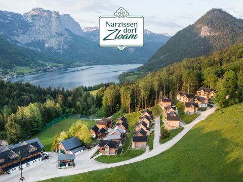 Luftaufnahme eines Dorfes mit Holzhütten, umgeben von üppigem Grün und Wäldern, in der Nähe eines Sees und der Berge. Oben steht ein Schild mit der Aufschrift „Narzissendorf Zloam“. Die Szenerie ist friedlich und malerisch.