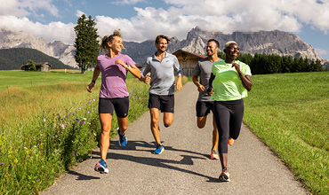 Vier Personen joggen auf einem Pfad durch eine üppig grüne Wiese mit Bergen und Wolken im Hintergrund. Sie tragen sportliche Kleidung und lächeln, während sie gemeinsam laufen.