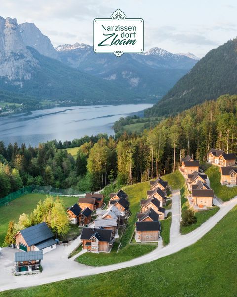 Luftaufnahme des Narzissendorfs Zloam. Sie zeigt eine Ansammlung rustikaler Häuser, eingebettet in üppiges Grün nahe einem ruhigen See. Die Berge im Hintergrund runden die malerische Szenerie ab.