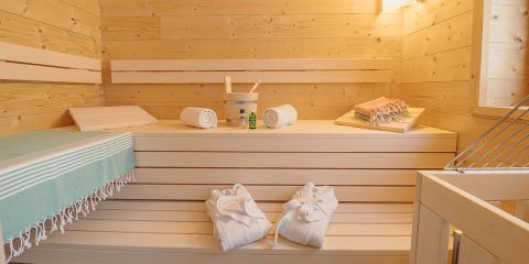 Ein Sauna-Innenraum aus Holz mit abgestuften Bänken, ordentlich platzierten Handtüchern und einem Eimer mit Schöpfkelle. Ein Paar weiße Hausschuhe liegt auf dem Boden, und gefaltete Handtücher mit Flasche und Bürste liegen auf einer Bank und schaffen so eine gemütliche Spa-Atmosphäre.
