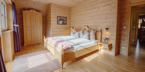 Ein gemütliches Schlafzimmer mit Holzwänden und -boden verfügt über ein Doppelbett mit geblümter Bettwäsche, einen Holzschrank und einen kleinen Nachttisch mit Blumen. Ein Fenster mit lila Vorhängen lässt Tageslicht herein, und an der Wand hängt ein gerahmtes Bild.